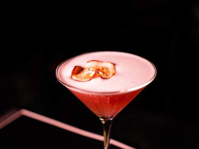 En lyserød cocktail i et martini-glas med en skummende top, pyntet med to tynde æbleskiver, på en mørk baggrund.