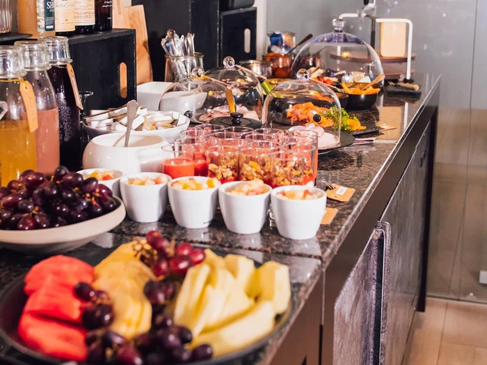 Et buffetbord med forskelligt frugt, granola-parfaits, juice, pålæg, ost og overdækkede fade på en mørk bordplade.