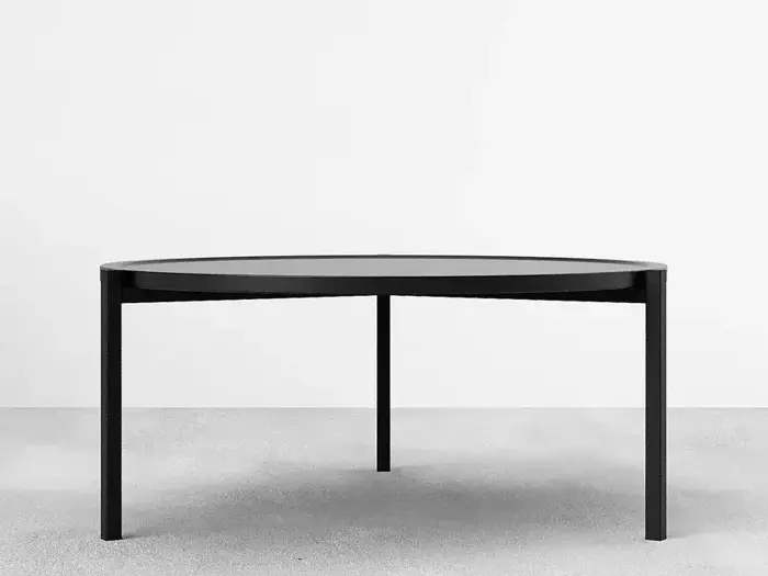 Minimalistisk rundt sort bord med tre ben, placeret på et lysegråt gulv mod en almindelig hvid baggrund.