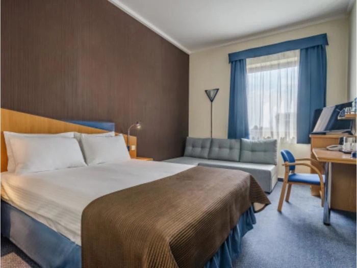 Et hotelværelse med dobbeltseng, brunt tæppe, grå sofa, skrivebord med stol, lampe og et vindue med blå gardiner, der lukker dagslyset ind.