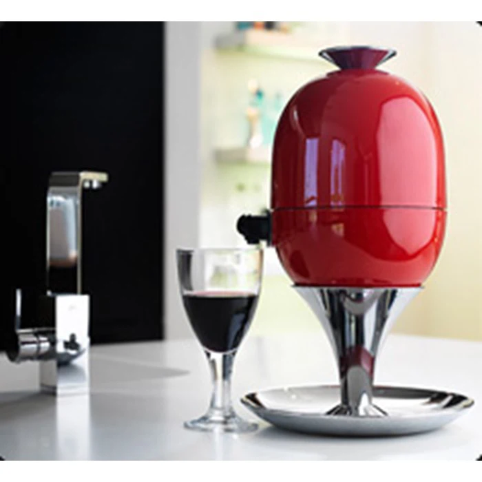 En moderne rødvinsdispenser formet som et æg står på en kromfod ved siden af et glas rødvin på en hvid bordplade.