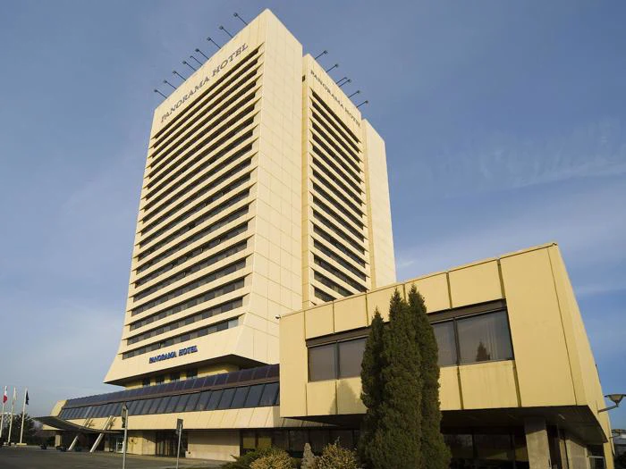 Høj gul-hvid hotelbygning med National Hotel-skilt, store vinduer og træer foran mod en klar blå himmel.