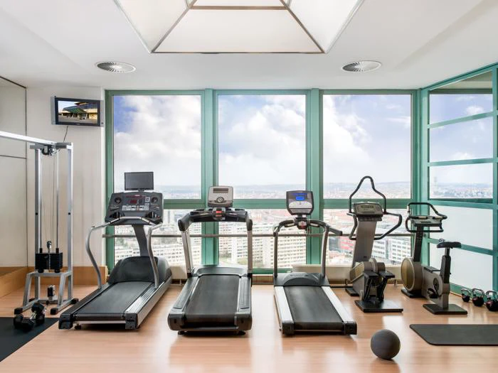 Moderne fitnesscenter med store vinduer, løbebånd, motionscykler, en romaskine, vægte og en kabelmaskine med udsigt over et bybillede under en delvis overskyet himmel.