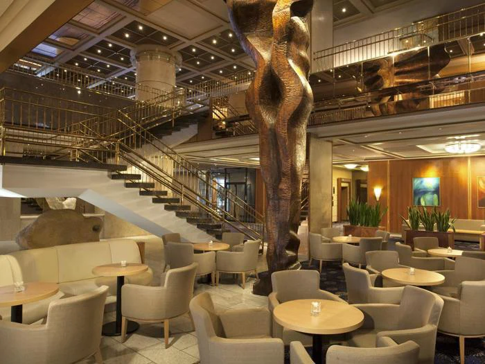 Moderne hotellounge med beige lænestole, runde borde, en stor abstrakt bronzeskulptur, stor trappe, planter og varm belysning. Øvre mezzanin med guldgelænder i baggrunden.