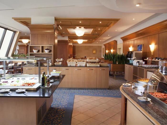 Hotellets morgenmadsbuffet med forskellige fødevarer, drikkevarer og kager på diske, pænt arrangeret i et veloplyst, moderne spiseområde med træindretning og blåt gulvtæppe.