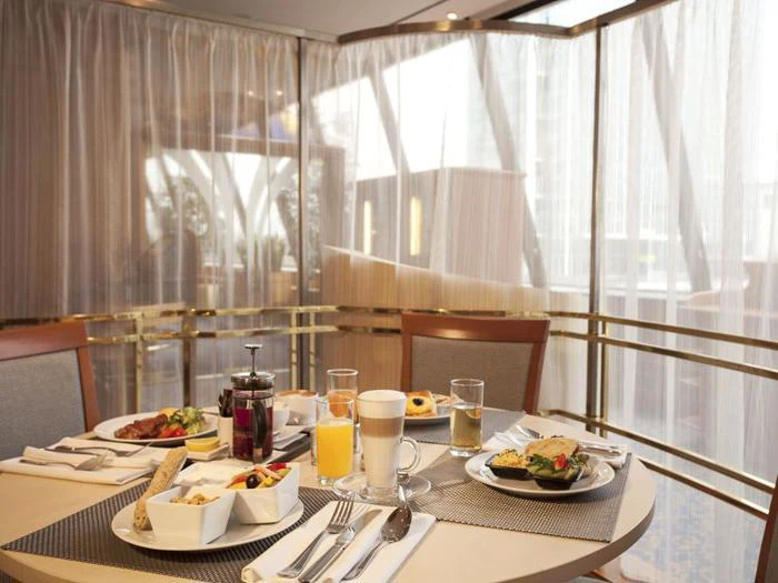 Et rundt morgenbord med juice, kaffe, frugt, kager og bestik er placeret ved store vinduer med gennemsigtige gardiner i et lyst, moderne spiseområde.