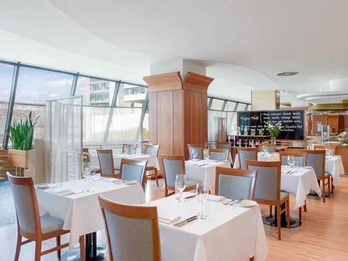 En moderne restaurant med træstole, hvide duge, pænt dækkede borde, store vinduer og et tavlemenu i nærheden af baren.