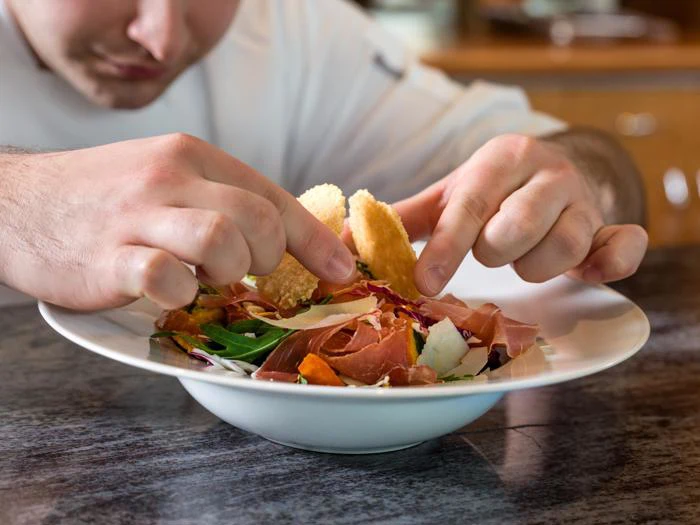 En kok arrangerer omhyggeligt ingredienser, herunder saltet kød og grønt, på et salatfad og fokuserer intenst på præsentationen.