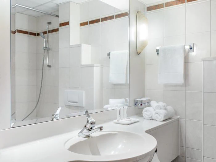 Moderne badeværelse med hvid håndvask, vandhane, stort spejl, håndklædeholder med hvide håndklæder, bruseområde og toiletartikler, der er arrangeret pænt på disken.