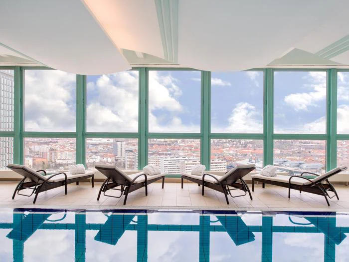 Indendørs pool med liggestole, der vender ud mod gulv-til-loft-vinduer med panoramaudsigt over byen under en delvis overskyet himmel. Håndklæder er pænt placeret på hver stol.