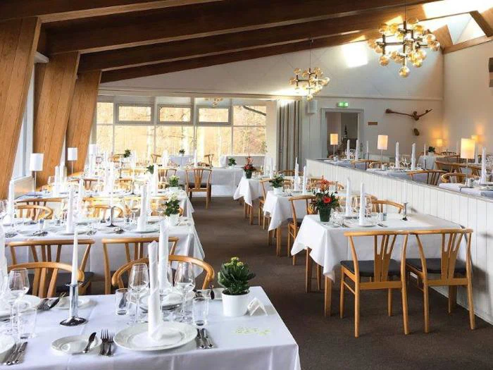 En lys, elegant restaurant med træbjælker, hvide duge, høje stearinlys og dekorative planter på sirligt opstillede borde. Store vinduer lukker naturligt lys ind.