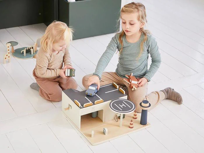 To små børn leger på gulvet med et parkeringshus i træ, biler og vejskilte i et lyst, hvidt rum.
