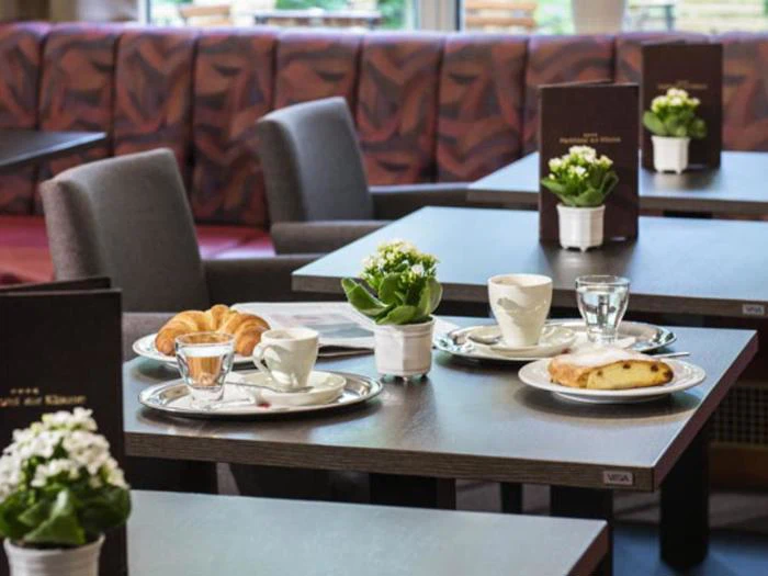 Et cafébord dækket med kaffekopper, en croissant, et stykke kage, vandglas, hvide blomster i potter og menukort, med flere borde og stole i baggrunden.