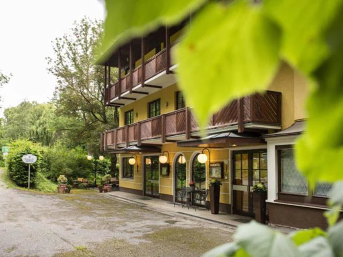 En gul bygning med træbalkoner og udendørslamper, delvis skjult af grønne blade, ligger ved siden af en stille vej med træer.