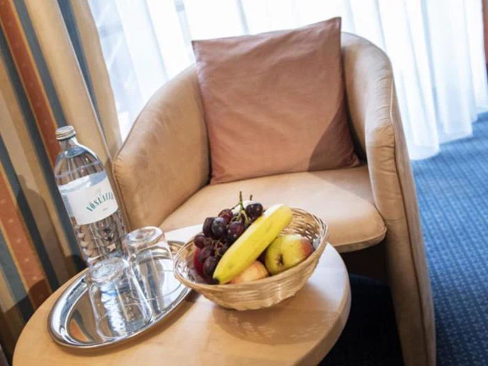 En beige lænestol med en lyserød pude står ved siden af et rundt bord med en frugtkurv, en flaske vand og to glas på en metalbakke. Der er lyse gardiner i baggrunden.