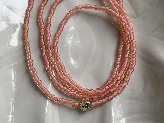 En pink perlehalskæde med små firkantede perler og en enkelt guldaccentperle, viklet på en hvid, blank overflade.