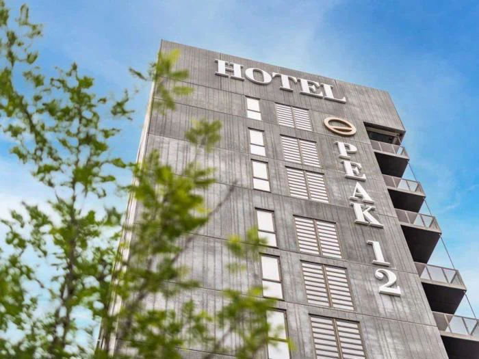 Høj moderne bygning med HOTEL PEAK 12 skrevet på facaden, fotograferet nedefra med en klar blå himmel og grønne trægrene i forgrunden.