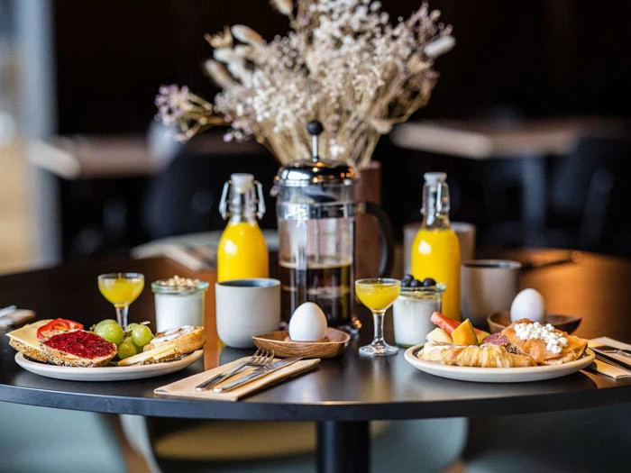 Et morgenbord med kager, toast, frugt, kogte æg, juice, en fransk presse, kaffekopper og en vase med tørrede blomster i midten.