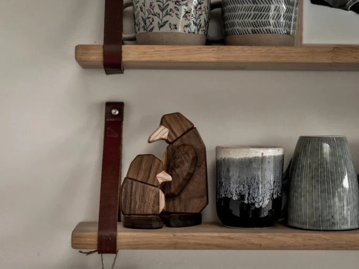 Pingvinfigurer af træ på en træhylde ved siden af keramikkrus og kopper, med en læderstrop, der støtter hylden.
