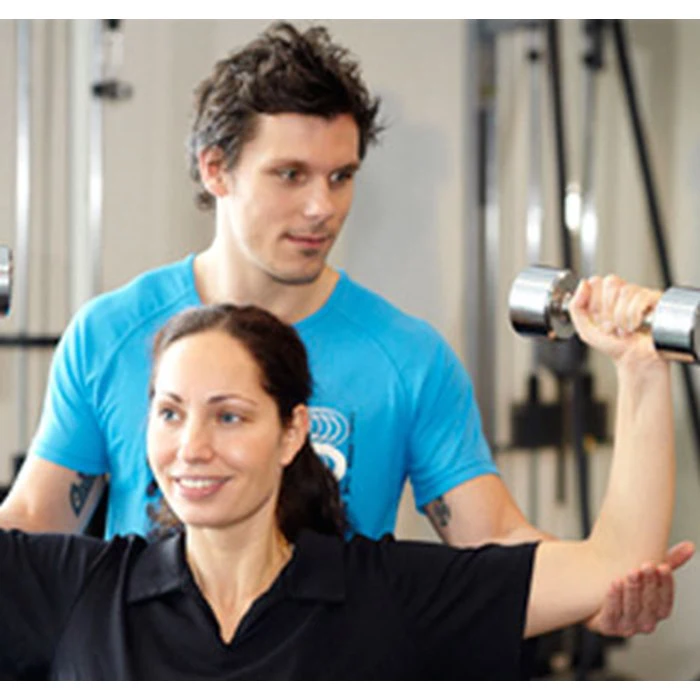En kvinde løfter håndvægte, mens en mandlig træner i blå skjorte hjælper hende i fitnesscentret.