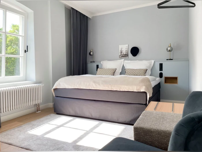 Moderne soveværelse med en stor seng, to puder, væglamper, et lille indrammet billede, et lyst tæppe, et vindue med sollys, en radiator og minimalistisk indretning.