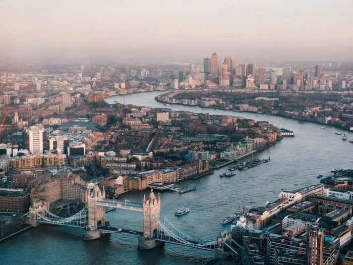 Luftfoto af London med Tower Bridge over Themsen, med byens bygninger og Canary Wharf skyline i det fjerne.
