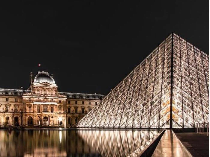 Louvre-museets oplyste glaspyramide står ved siden af den historiske bygning om aftenen og spejler sig i vandet nedenunder.