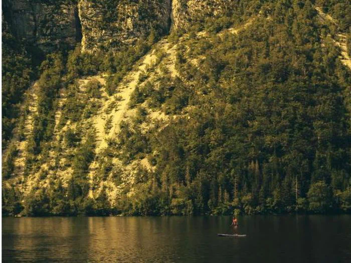 En person på paddleboard på en rolig sø med en stejl, træbevokset bjergskråning og klipper i baggrunden.