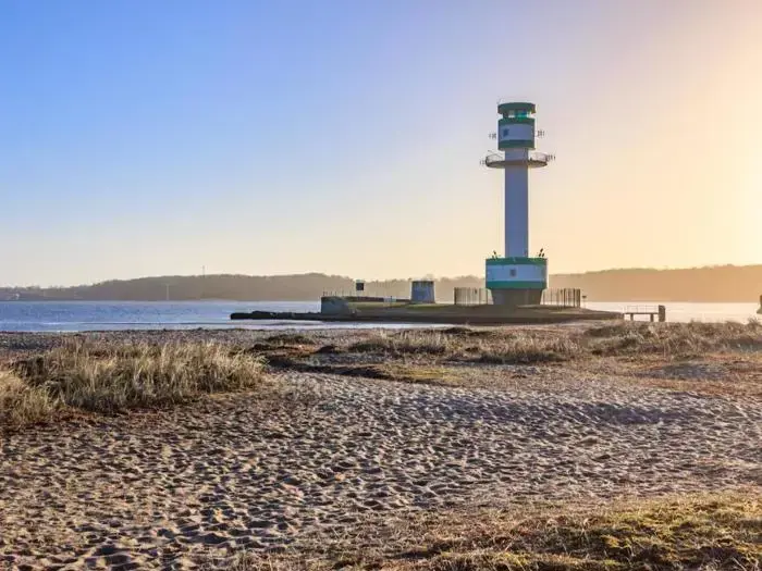 Et fyrtårn står på en mole ved vandet med sandstrand og tørt græs i forgrunden under en klar blå himmel ved solnedgang.