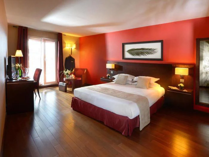 Et moderne hotelværelse med en stor seng, trægulve, røde accentvægge, et skrivebord, en stol, kunstværker over sengen og naturligt lys fra en terrassedør.