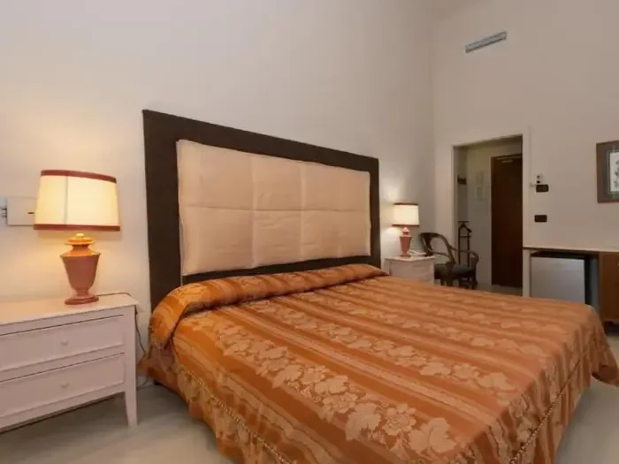 Et hotelværelse med en dobbeltseng dækket af et orangemønstret sengetæppe, to natborde med lamper, en stol, et lille køleskab og en dør i baggrunden.