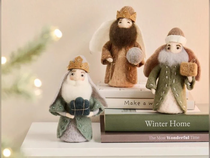 Tre figurer af filtet uld, der ligner de tre vise mænd, står på en stak bøger omgivet af julepynt i en hyggelig atmosfære.