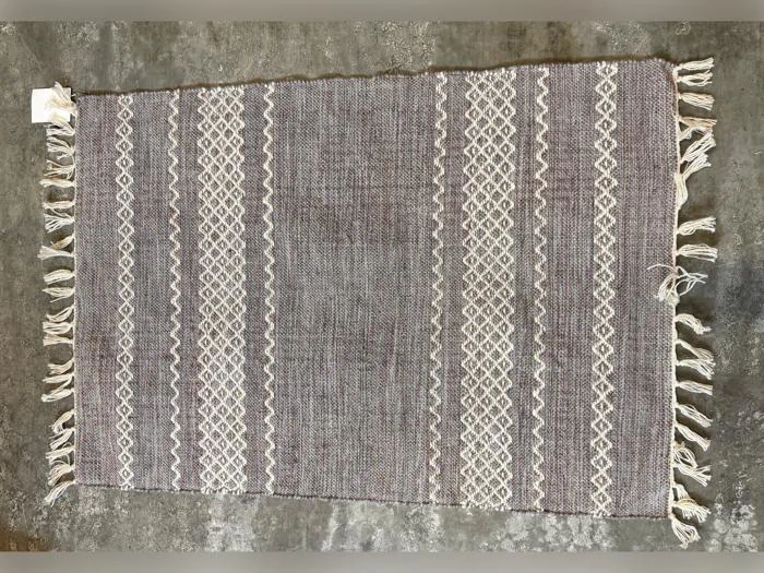 Vævet tæppe med beige og hvide geometriske striber, kvaster i de korte ender og en struktureret grå baggrund, placeret på et betongulv.
