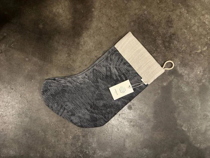 En mønstret julesok i antracitgrå med en lys kant og en hængemærkat på et mørkt betongulv.