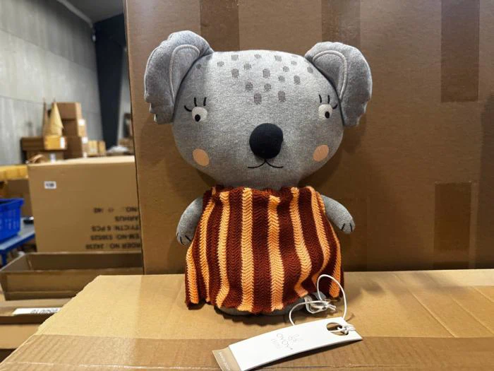 Plys-koala med et gråt mønstret hoved og en brun og orange stribet kjole, med en papirmærkat fastgjort.