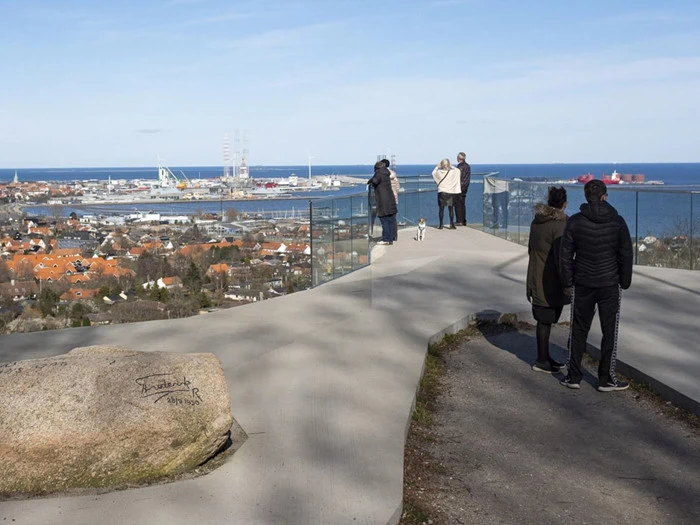 Flere mennesker står på et moderne observationsdæk af glas med udsigt over en kystby, havnen og havet under en delvis overskyet himmel. En hund er også til stede.