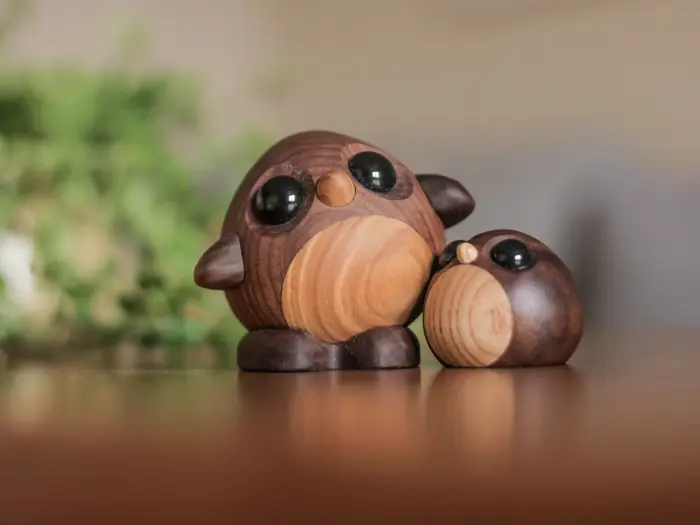 To små, runde uglefigurer af træ med store, sorte øjne er placeret på en glat overflade med en sløret, grøn plante i baggrunden.