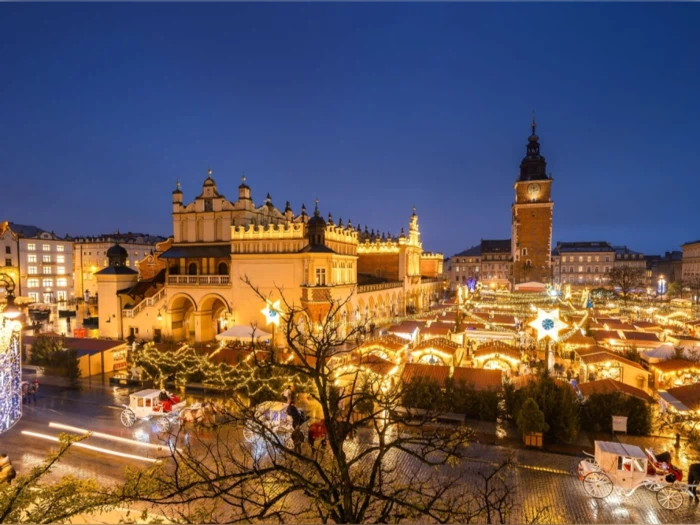 Et festligt julemarked lyser op på et europæisk torv om aftenen med historiske bygninger og et højt klokketårn.