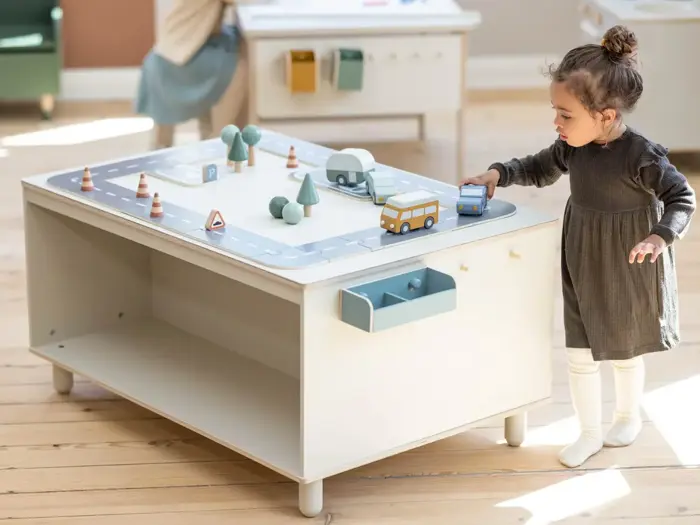 Et lille barn leger med legetøjskøretøjer på en bordplade med byveje, mens det står ved siden af et hvidt legebord i et lyst rum.