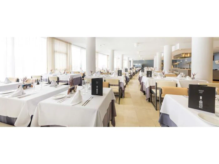 En lys, moderne restaurant med pænt opstillede borde med hvide duge, dækket med glas, servietter og menuer, omgivet af store vinduer og hvide søjler.