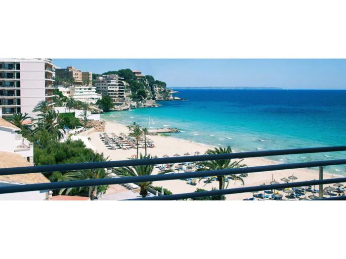 Udsigt over en sandstrand med parasoller, klart blåt hav, palmer og nærliggende hoteller og bygninger, set bag et balkonrækværk.