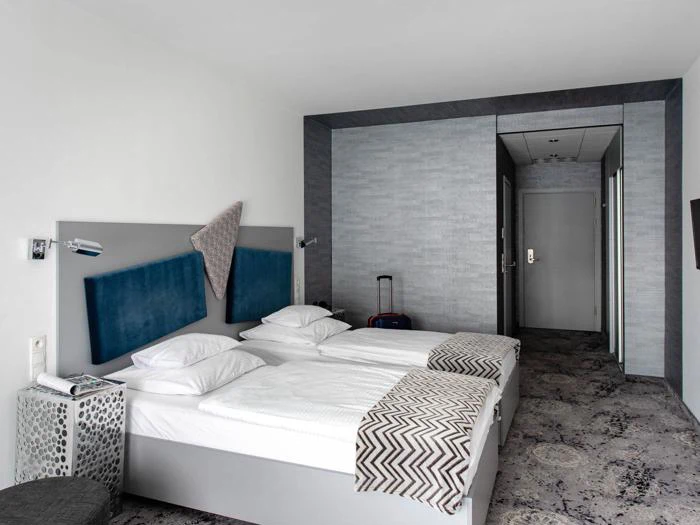 Moderne hotelværelse med to enkeltsenge skubbet sammen, hvidt sengetøj, blå sengegavle, mønstrede tæpper, et lille bord og en kuffert ved døren.