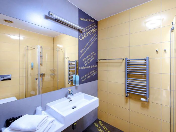 Moderne badeværelse med gule flisevægge, hvid håndvask, spejl, håndklædetørrer, brusekabine i glas og motiverende citater skrevet på væggen på italiensk.