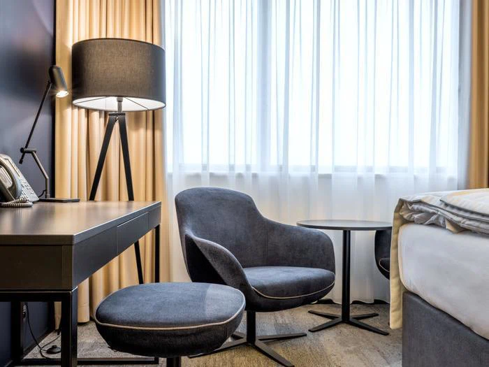 Moderne hotelværelseshjørne med en grå lænestol, matchende ottoman, lille rundt bord, gulvlampe, skrivebord og seng nær et stort vindue med gennemsigtige gardiner.