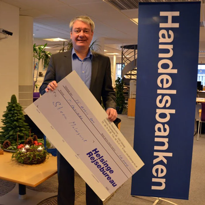 En smilende mand på et kontor holder en stor check fra Helsinge Rejsebureau ved siden af et blåt Handelsbanken-banner og et skrivebord med små juletræer.