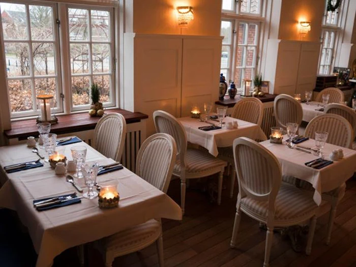 Elegant restaurant med hvide duge, stearinlys, glas og vintagestole ved siden af store vinduer, der lukker naturligt lys ind.