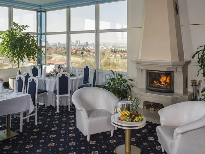 Et hyggeligt spiseområde med hvide stole, borde dækket til et måltid, en tændt pejs, frugt på et lille bord og store vinduer med udsigt over bybilledet.