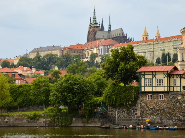 En udsigt over Prags borg og de omkringliggende historiske bygninger på en bakke med træer og en flod i forgrunden under en overskyet himmel.