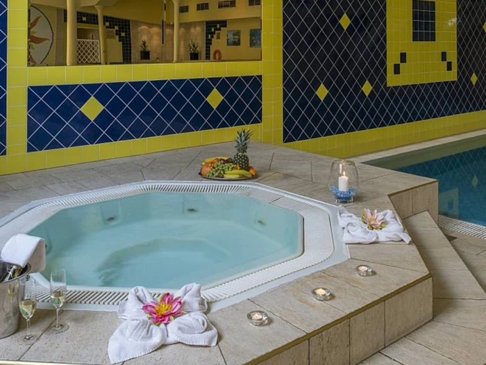 Indendørs spabad ved siden af en pool, omgivet af håndklæder, stearinlys, champagne, frugtfad og blomster; blå og gule flisevægge i baggrunden.