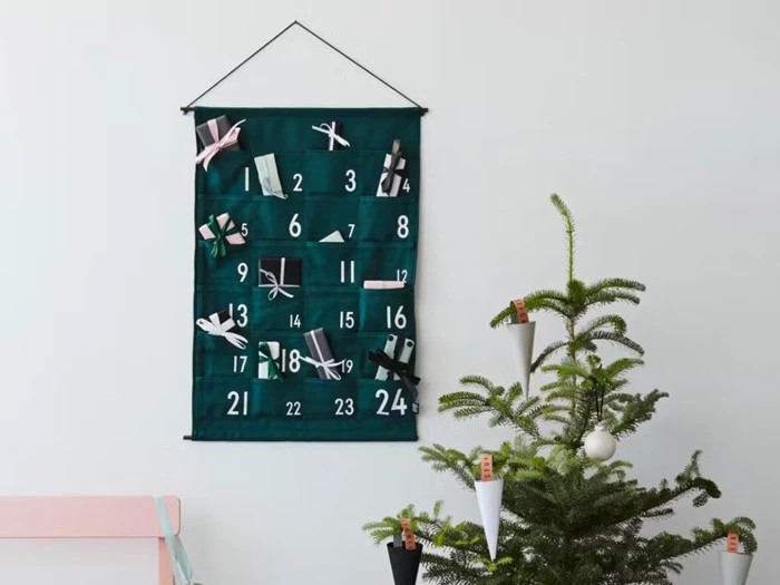 En grøn stofadventskalender med lommer nummereret 1-24 hænger på en væg over et pyntet juletræ med koglepynt.